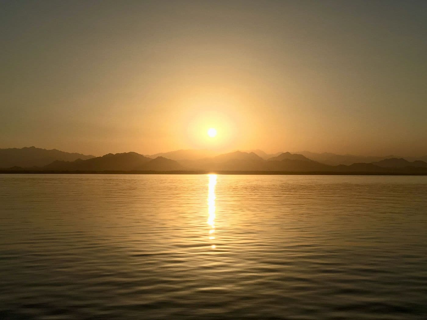 Sun low over a mountain range at dusk, its light reflected across still water